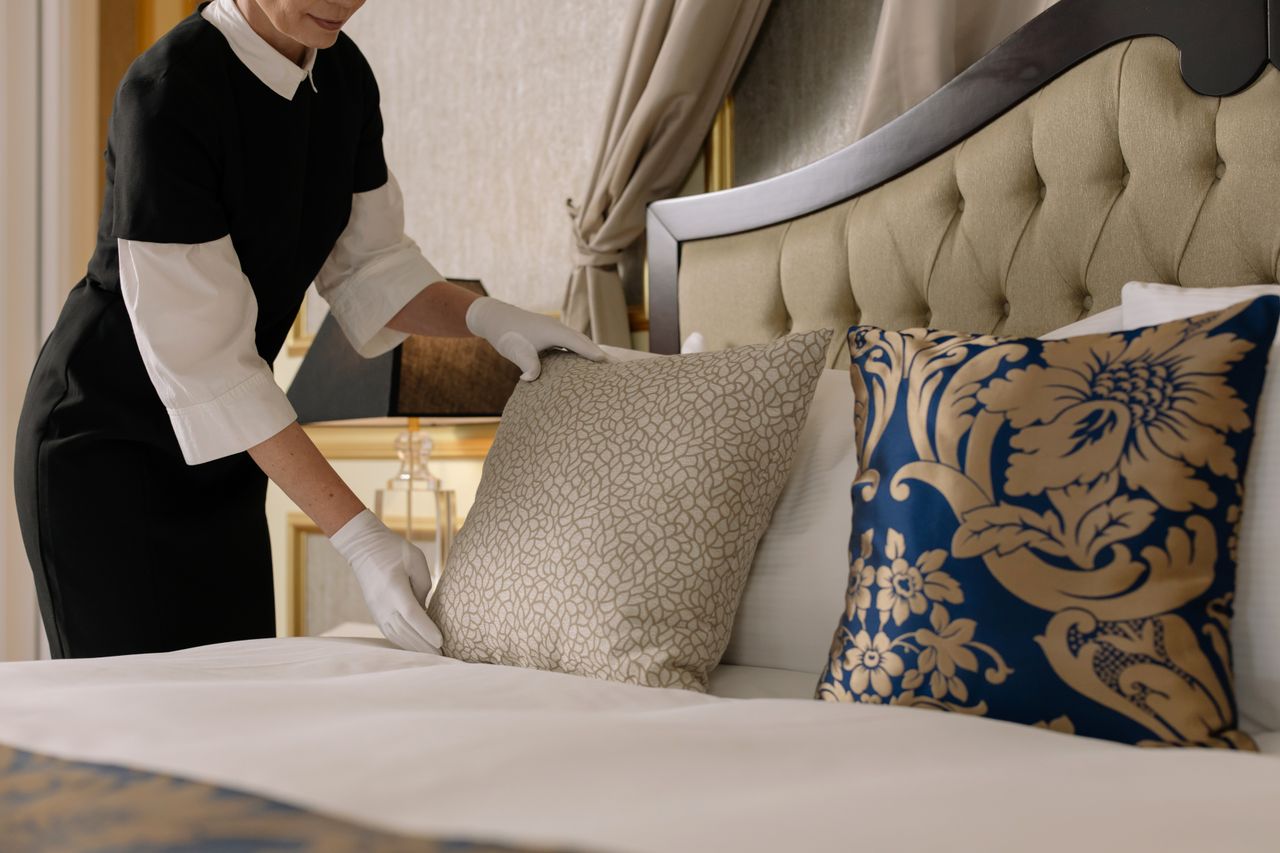 services-01 A chambermaid arranges pillows in a luxurious hotel room, showcasing comfort and elegance.