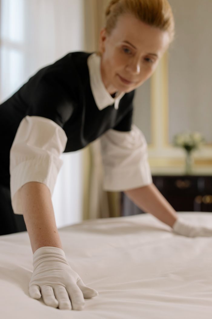 gallery-3 A housekeeper elegantly arranges bed sheets in a luxurious hotel room, showcasing hospitality expertise.